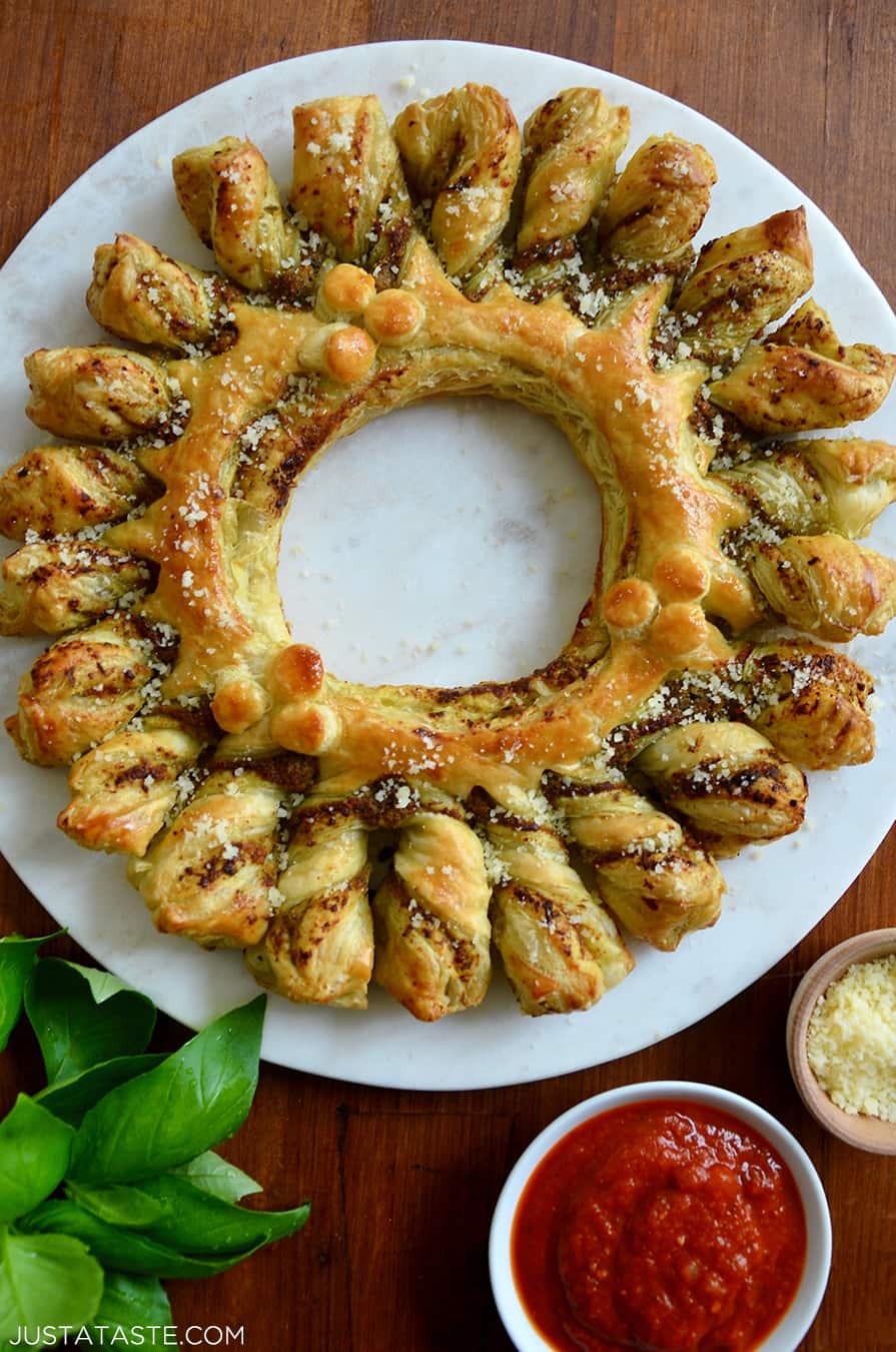 A top-down view of a pull-apart Pesto Puff Pastry Wreath next to fresh basil and small bowls containing marinara sauce and parmesan