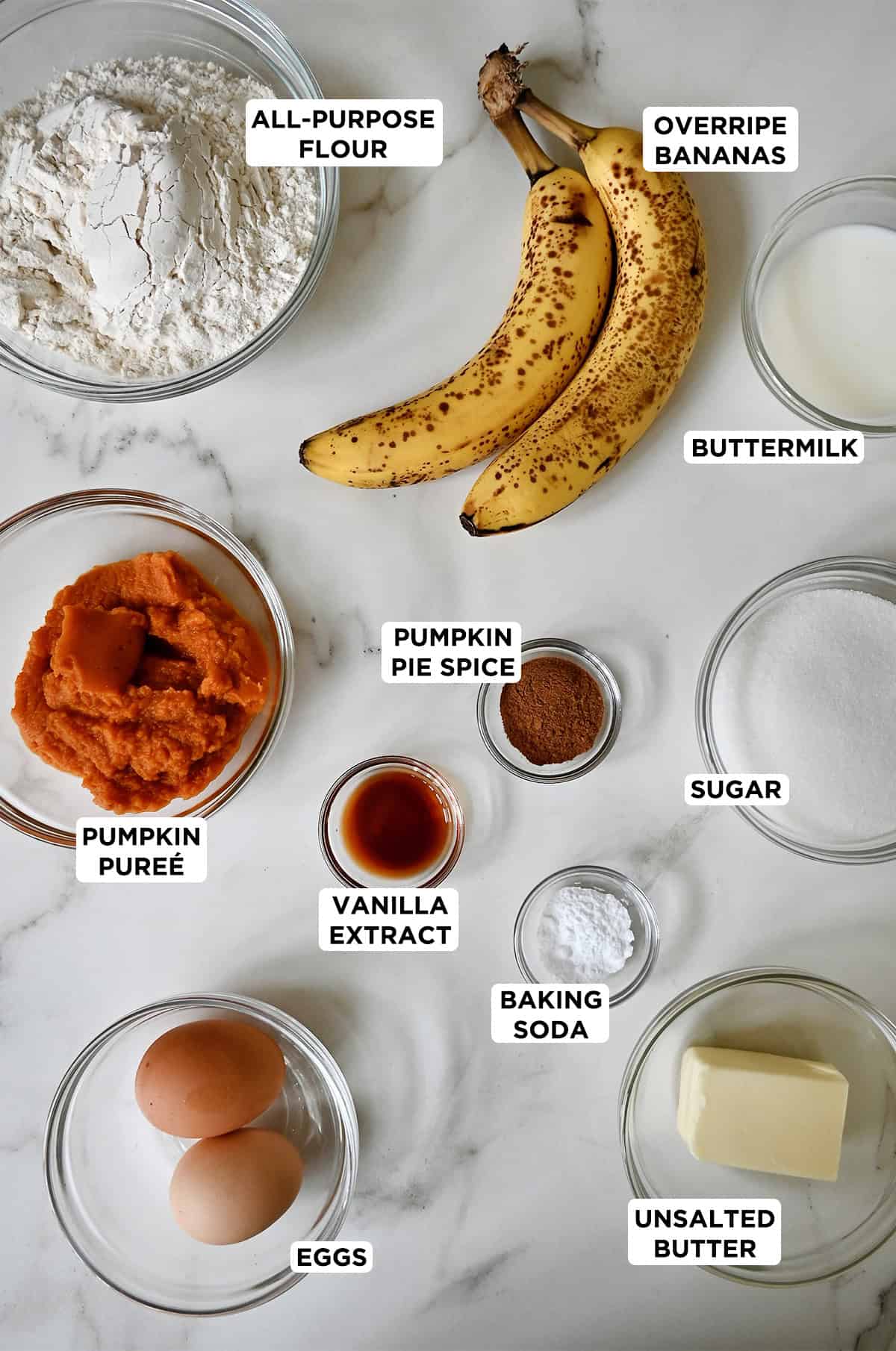 Ingredients to make pumpkin banana bread in glass bowls, including two overripe bananas, pumpkin purée, flour, eggs, spices, sugar, butter and buttermilk.
