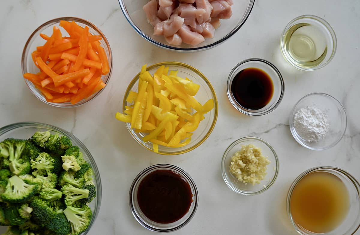 Glass bowls are filled with stir fry ingredients, including bell peppers, broccoli, soy sauce, raw chicken and minced garlic.