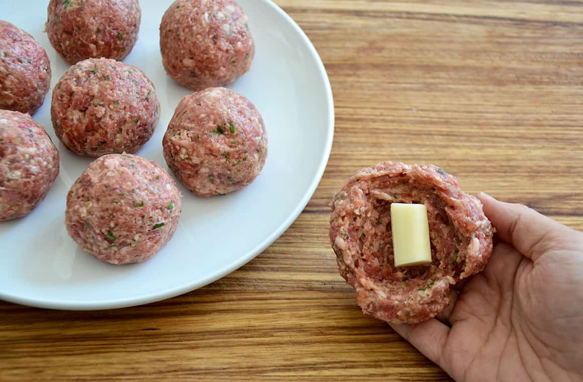 A hand holding an uncooked meatball with cheese in the center