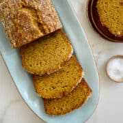 A half-sliced loaf of Pumpkin Banana Bread on a blue serving plate next to two overripe bananas and a small bowl filled with sanding sugar.