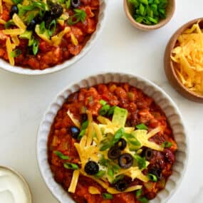 Two bowls containing leftover turkey chili topped with sour cream, shredded cheddar cheese, black olives and diced avocado.