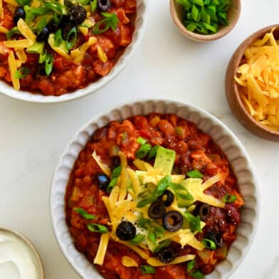 Two bowls containing leftover turkey chili topped with sour cream, shredded cheddar cheese, black olives and diced avocado.