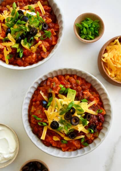 Two bowls containing leftover turkey chili topped with sour cream, shredded cheddar cheese, black olives and diced avocado.