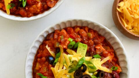 Two bowls containing leftover turkey chili topped with sour cream, shredded cheddar cheese, black olives and diced avocado.