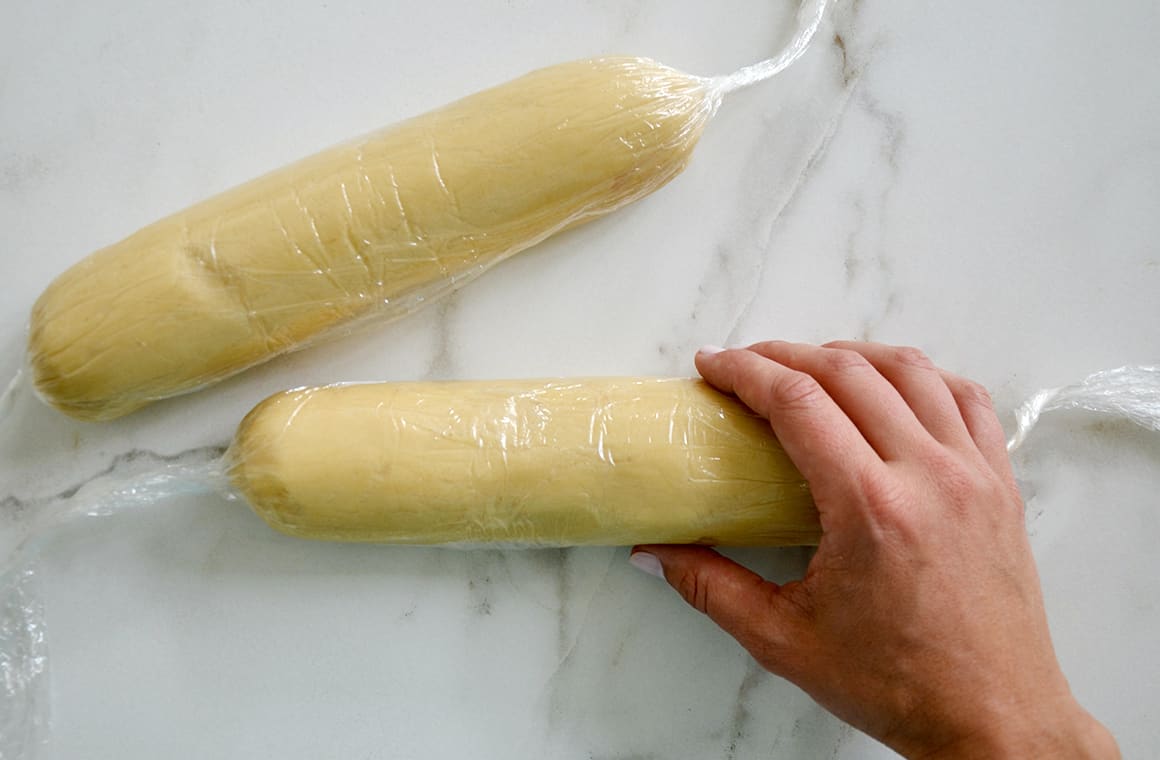 Two cookie dough logs wrapped in plastic wrap with a hand holding one