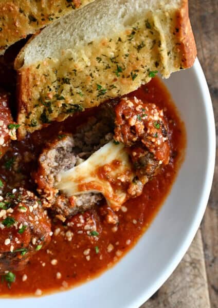 A bowl containing a gooey mozzarella stuffed meatball in marinara sauce and a slice of garlic bread for dipping.