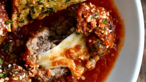 A bowl containing a gooey mozzarella stuffed meatball in marinara sauce and a slice of garlic bread for dipping.