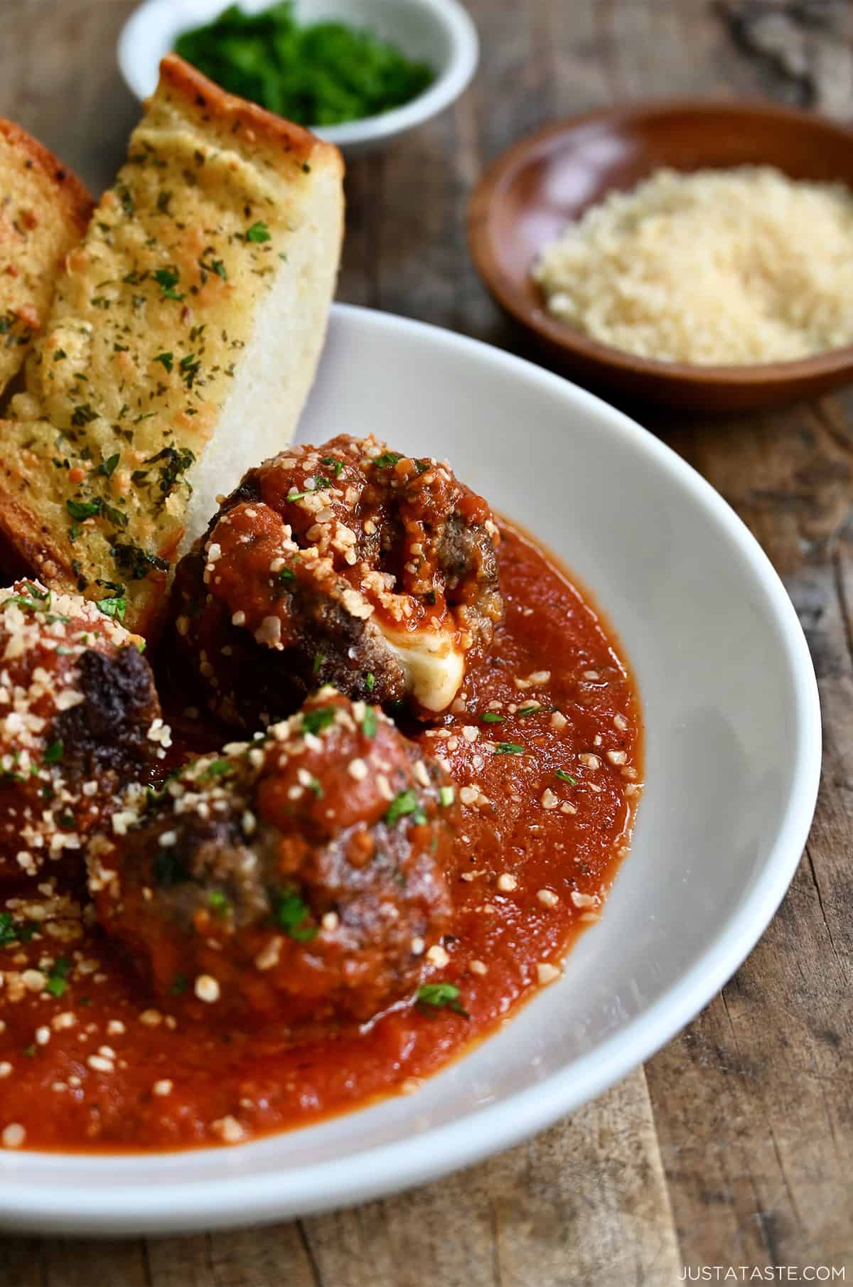 A bowl containing cheese stuffed Italian meatballs in marinara sauce. A slice of crusty garlic bread rests on the edge of the bowl.