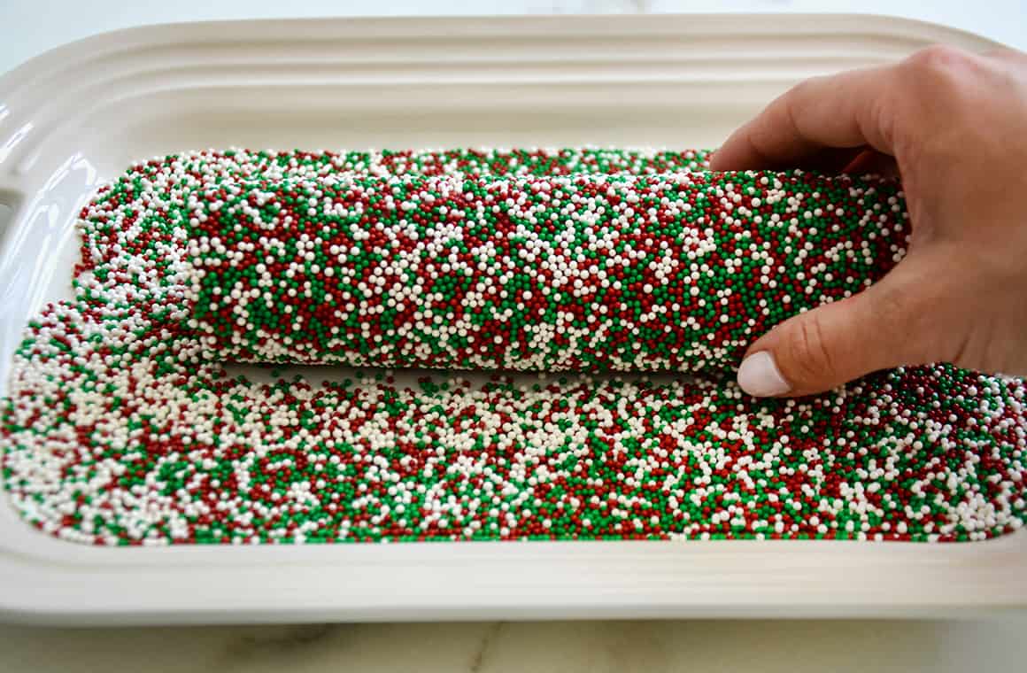 A hand rolling a cookie dough log in red, green and white sprinkles