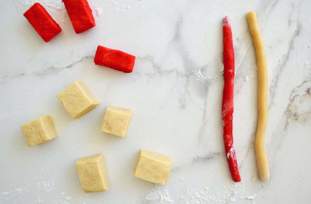 Red colored and plain cookie dough cubes and logs on a marble surface.