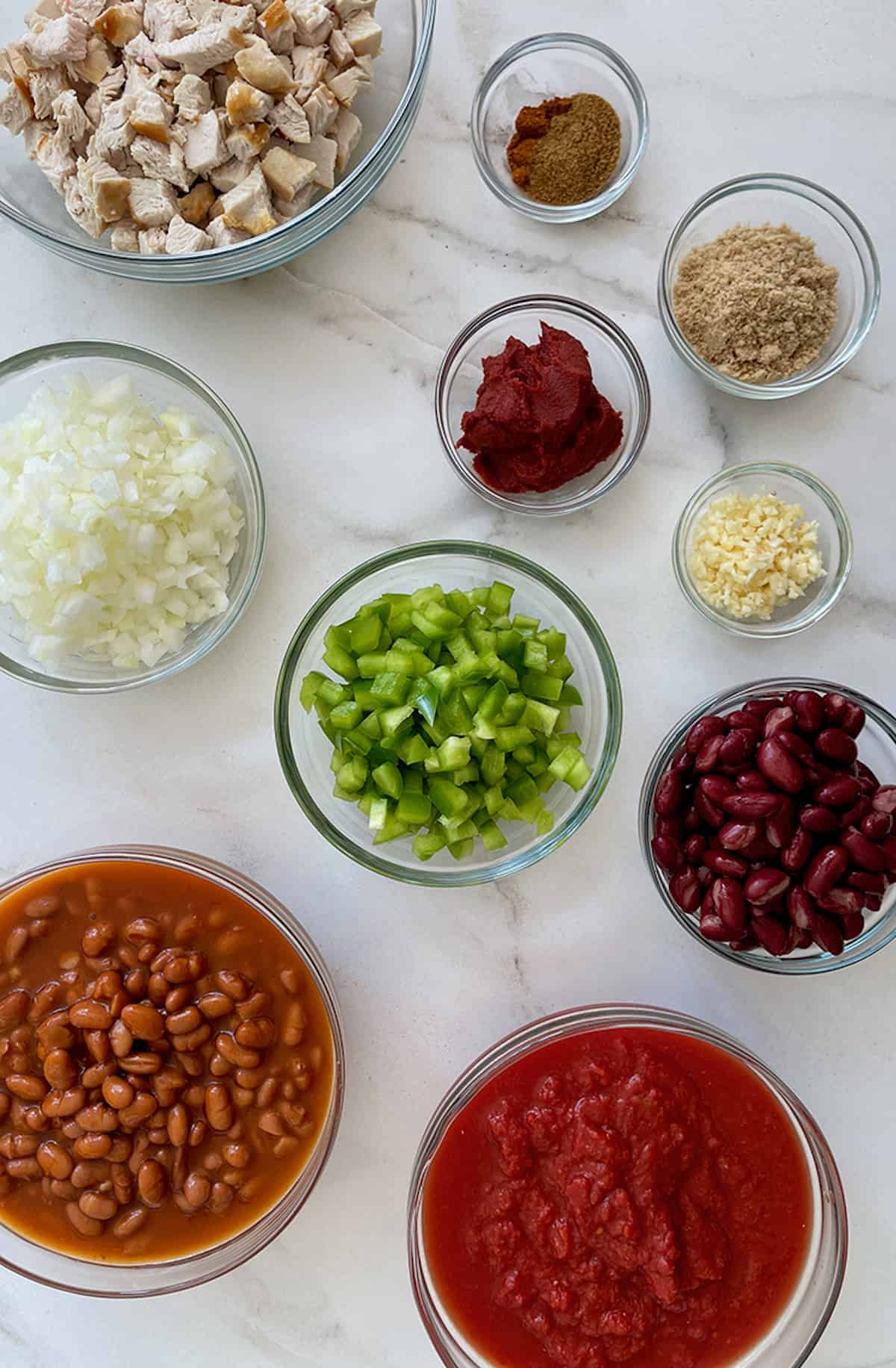 Various sizes of clear bowls containing chopped turkey, tomato paste, minced garlic, seasonings, brown sugar, diced green pepper, kidney beans, pinto beans, crushed tomatoes and diced onions.