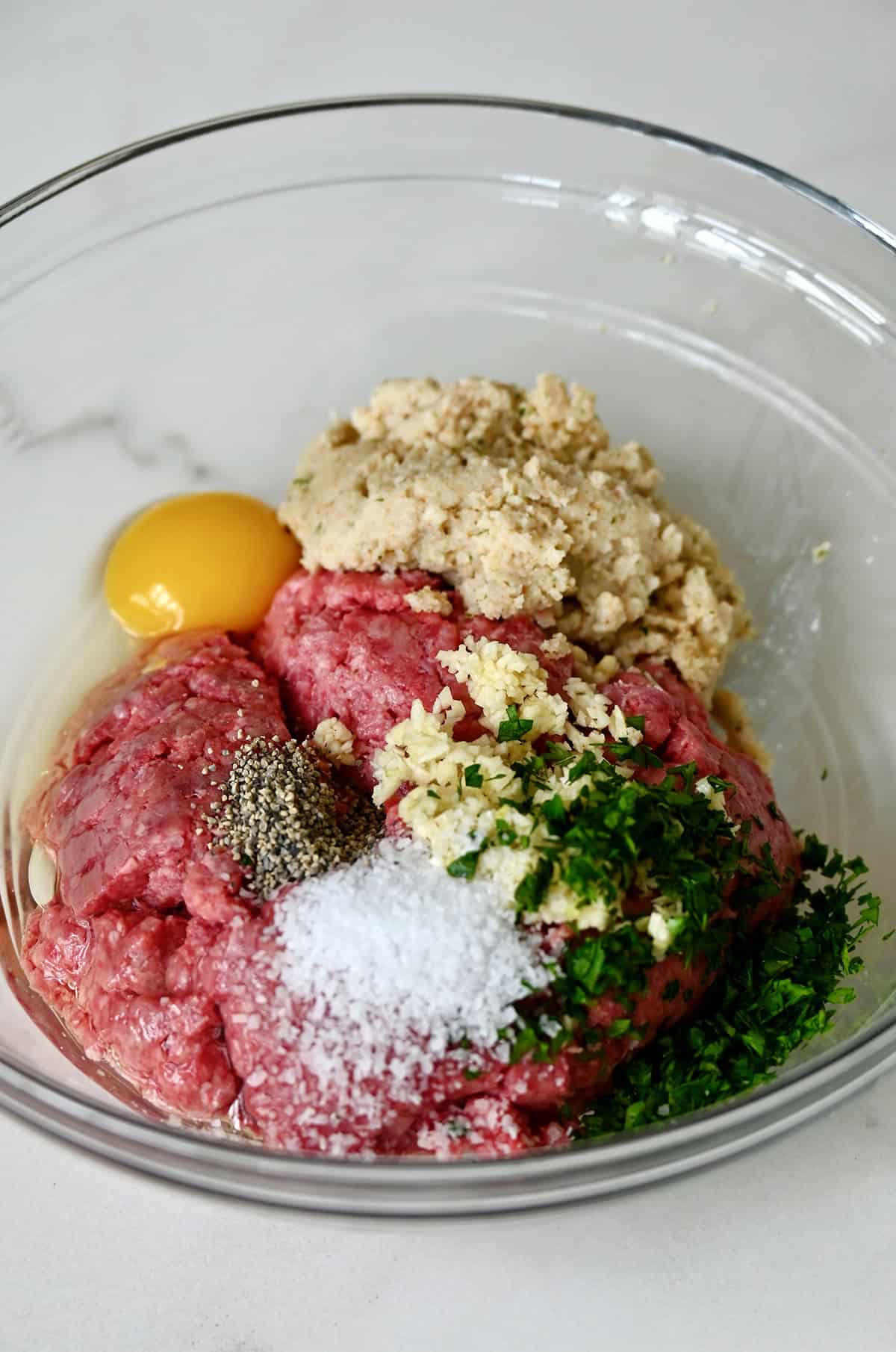 Ground meat in a bowl with an egg, panade, salt, pepper, minced garlic, and parsley. 