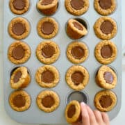 A child's hand reaching for a Mini Peanut Butter Cup Cookie in a muffin tin.