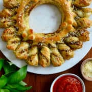 A top-down view of a pull-apart Pesto Puff Pastry Wreath next to fresh basil and small bowls containing marinara sauce and parmesan.