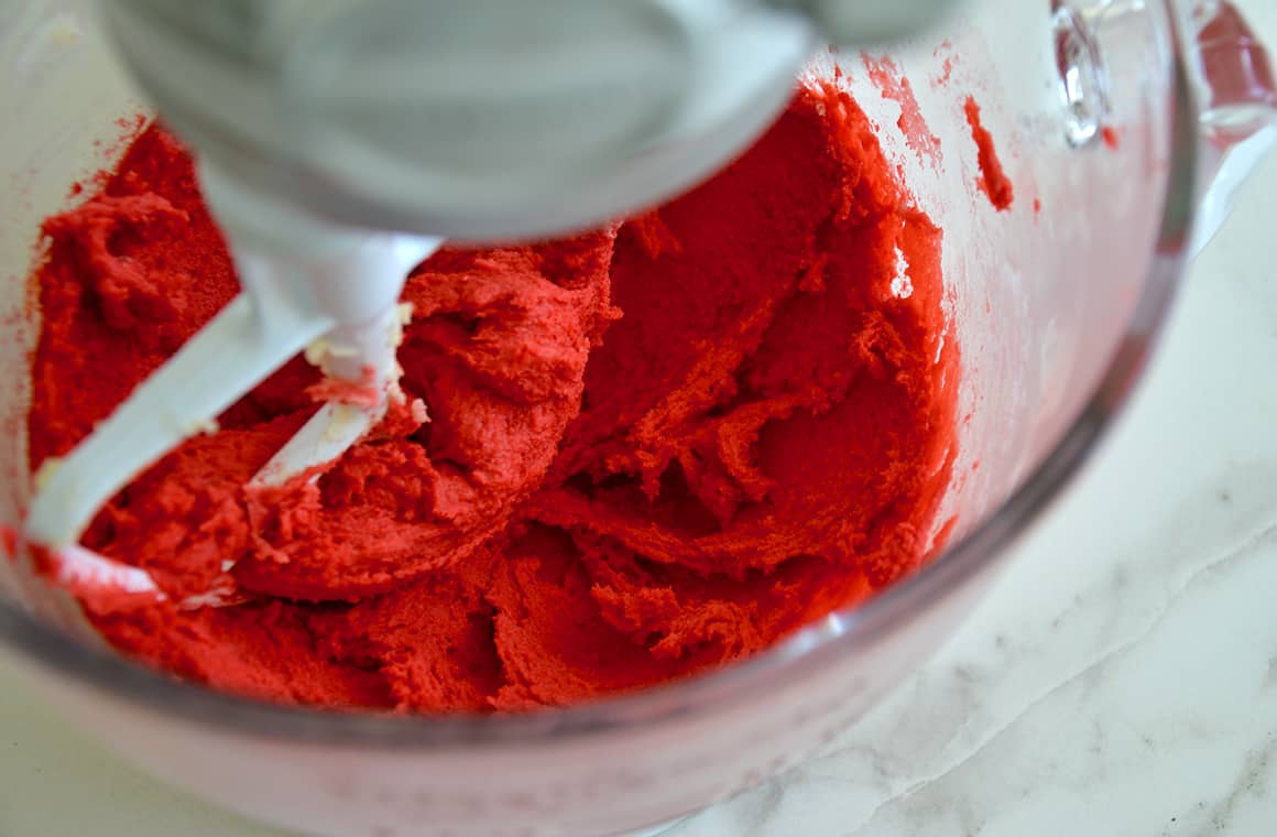 Red cookie dough in a stand mixer bowl with a paddle attachment