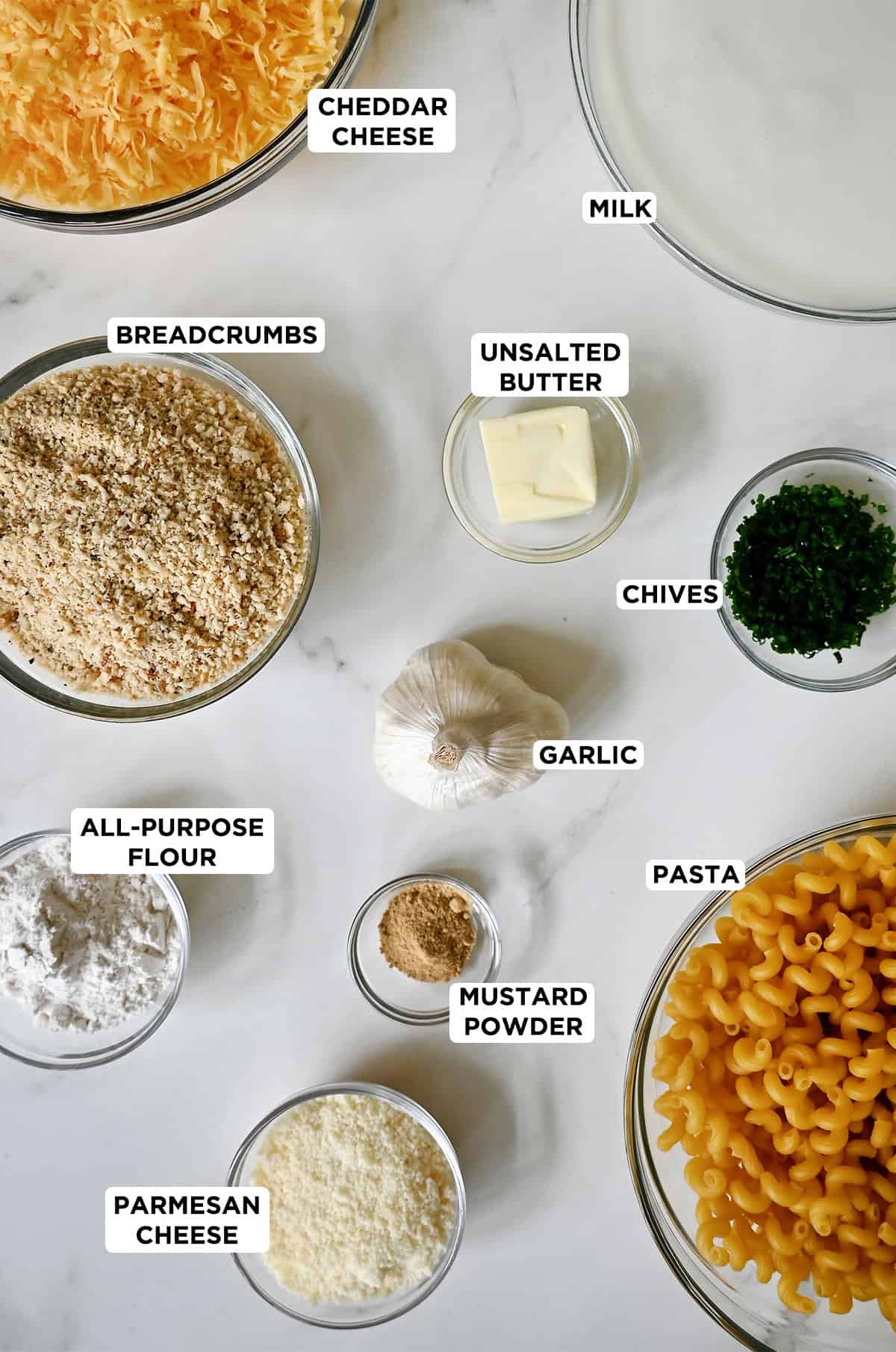 Various sizes of glass bowls containing ingredients to make baked macaroni and cheese with roasted garlic and a Panko breadcrumb topping.