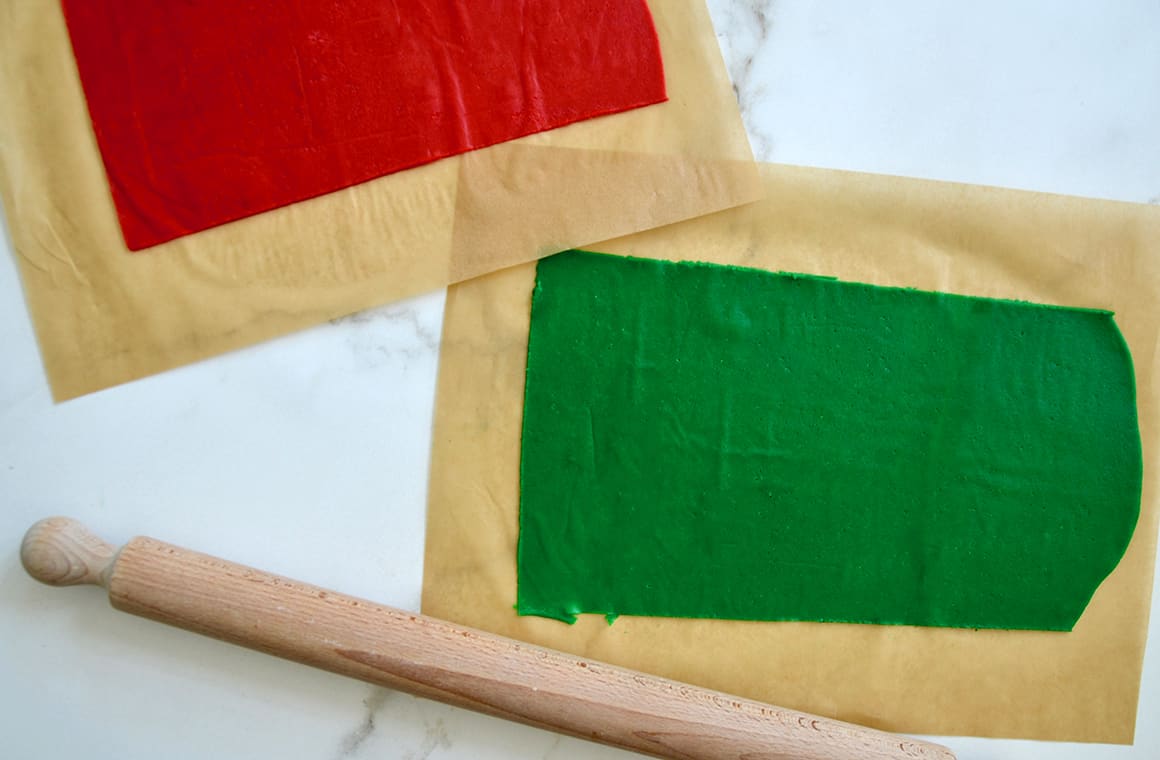 A rolling pin next to two pieces of parchment paper, one with red dough, the other with green 