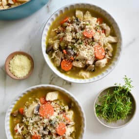 Two bowls containing homemade Chicken Wild Rice Soup next to small bowls containing parmesan and fresh herbs