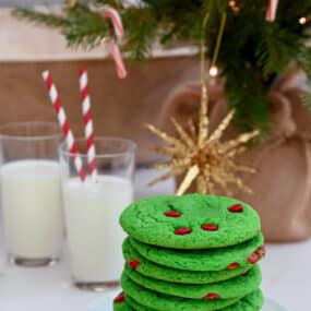 Green cake mix cookies with red M&Ms are stacked in front of glasses of milk with red striped straws. A Christmas tree is in the background.