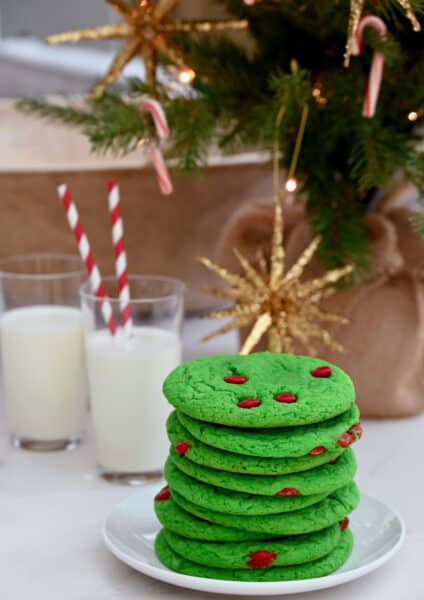 Green cake mix cookies with red M&Ms are stacked in front of glasses of milk with red striped straws. A Christmas tree is in the background.