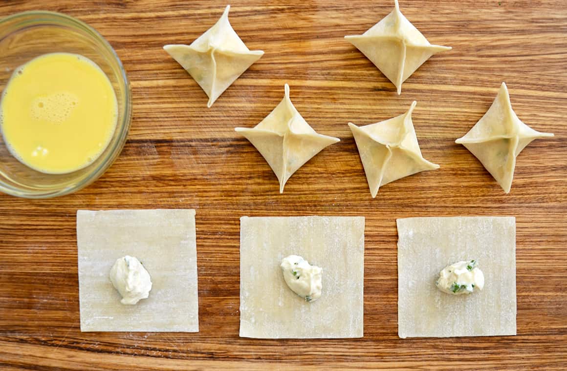 A top-down view of wonton wrappers topped with cream cheese mixture next to a small bowl containing egg wash mixture