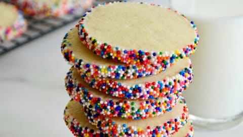 A stack of butter cookies with rainbow sprinkles next to a glass of milk and cookies cooling on a wire baking rack.
