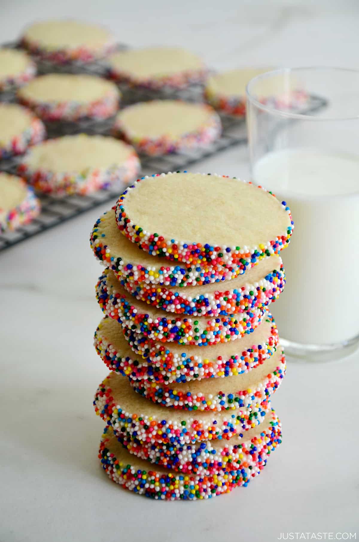 A stack of butter cookies with rainbow sprinkles next to a glass of milk and cookies cooling on a wire baking rack.