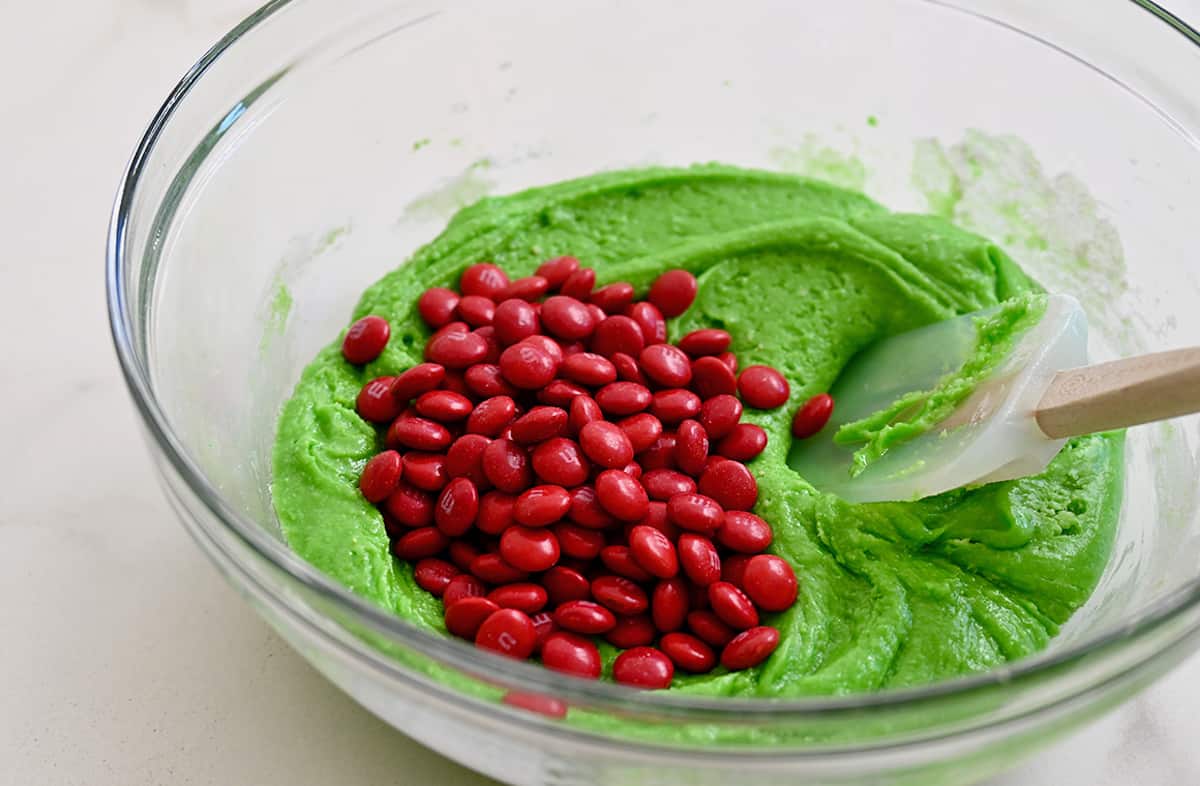 A glass bowl holds bright-green cake mix batter with red M&Ms ready to be stirred in with a spatula.