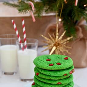 A tall stack of Cake Mix Grinch Cookies on a plate in front of two glasses of milk with straws