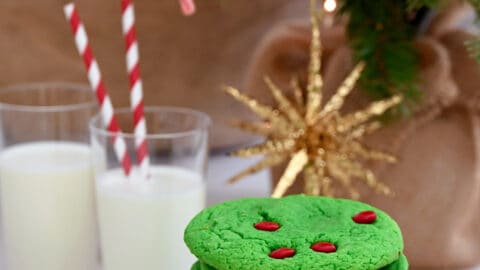 A tall stack of Cake Mix Grinch Cookies on a plate in front of two glasses of milk with straws