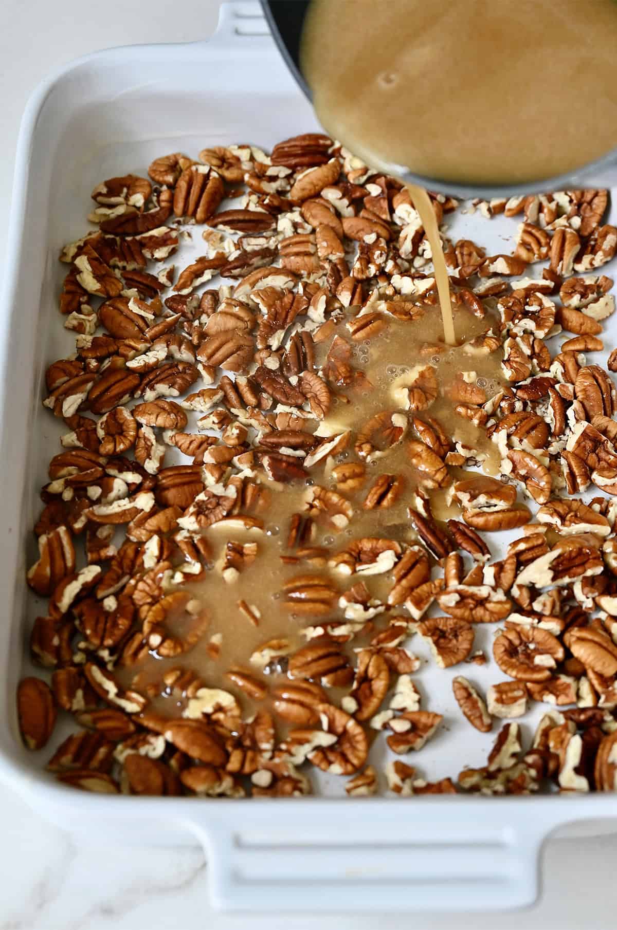 Pouring homemade caramel sauce over pecans in a baking dish.