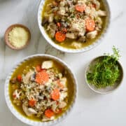 A top-down view of two bowls containing Chicken Wild Rice Soup next to two smaller bowls with fresh thyme and grated parmesan cheese and a blue stockpot containing soup.