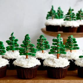 Two rows of chocolate cupcakes topped with cream cheese frosting, shredded coconut and pretzel candy melt Christmas tree toppers.