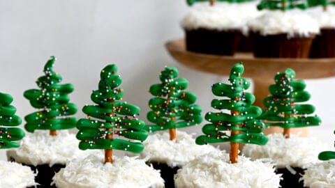Two rows of chocolate cupcakes topped with cream cheese frosting, shredded coconut and pretzel candy melt Christmas tree toppers.