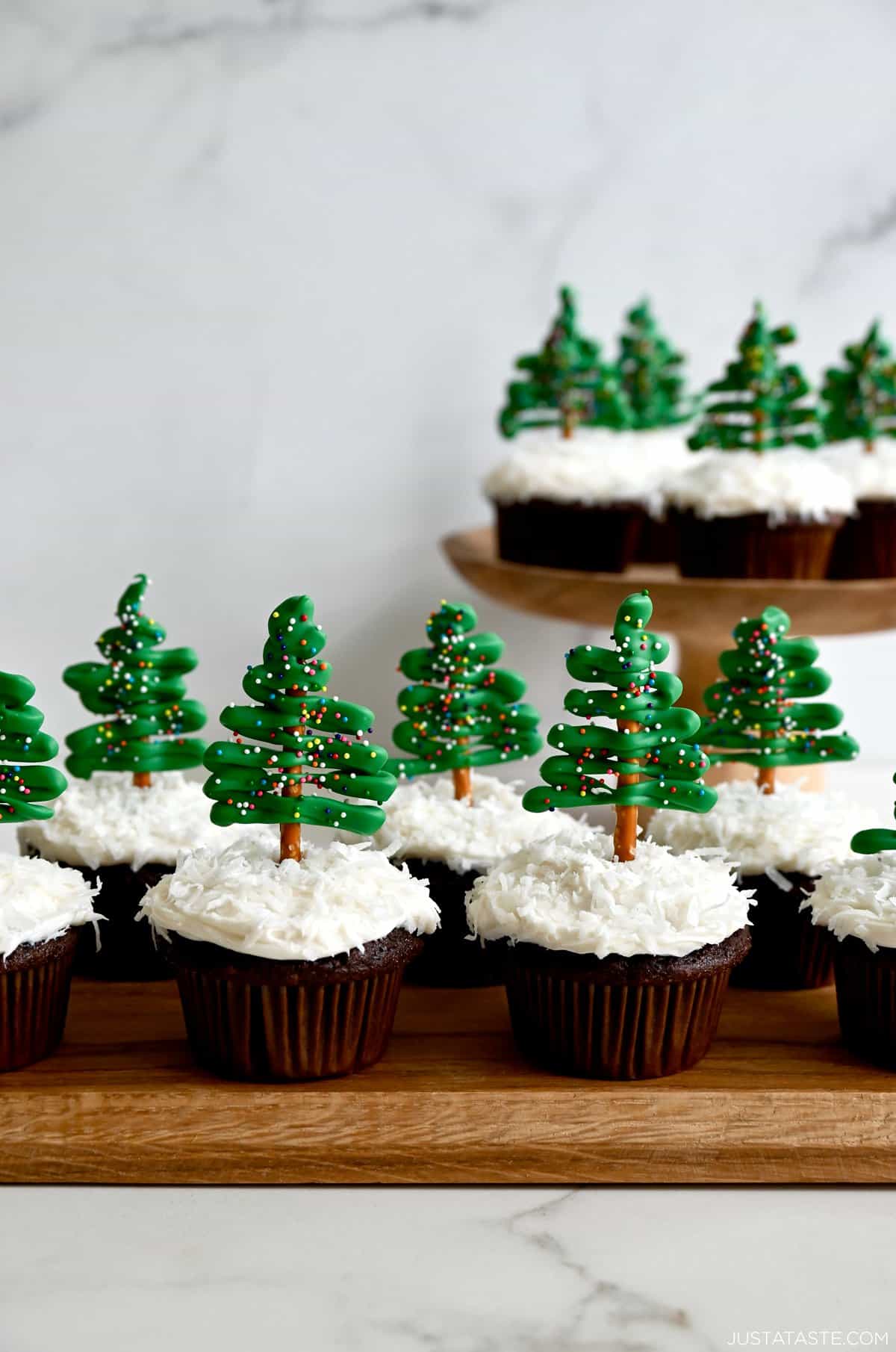 Two rows of chocolate cupcakes topped with cream cheese frosting, shredded coconut and pretzel candy melt Christmas tree toppers.