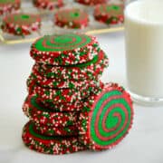 A tall stack of Christmas Pinwheel Cookies (red and green cookies) studded with festive sprinkles next to a tall glass of milk.