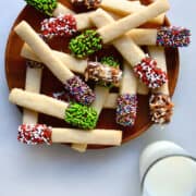 A stack of Sugar Cookie Sticks dipped in chocolate and studded with sprinkles on a serving plate next to a glass of milk.