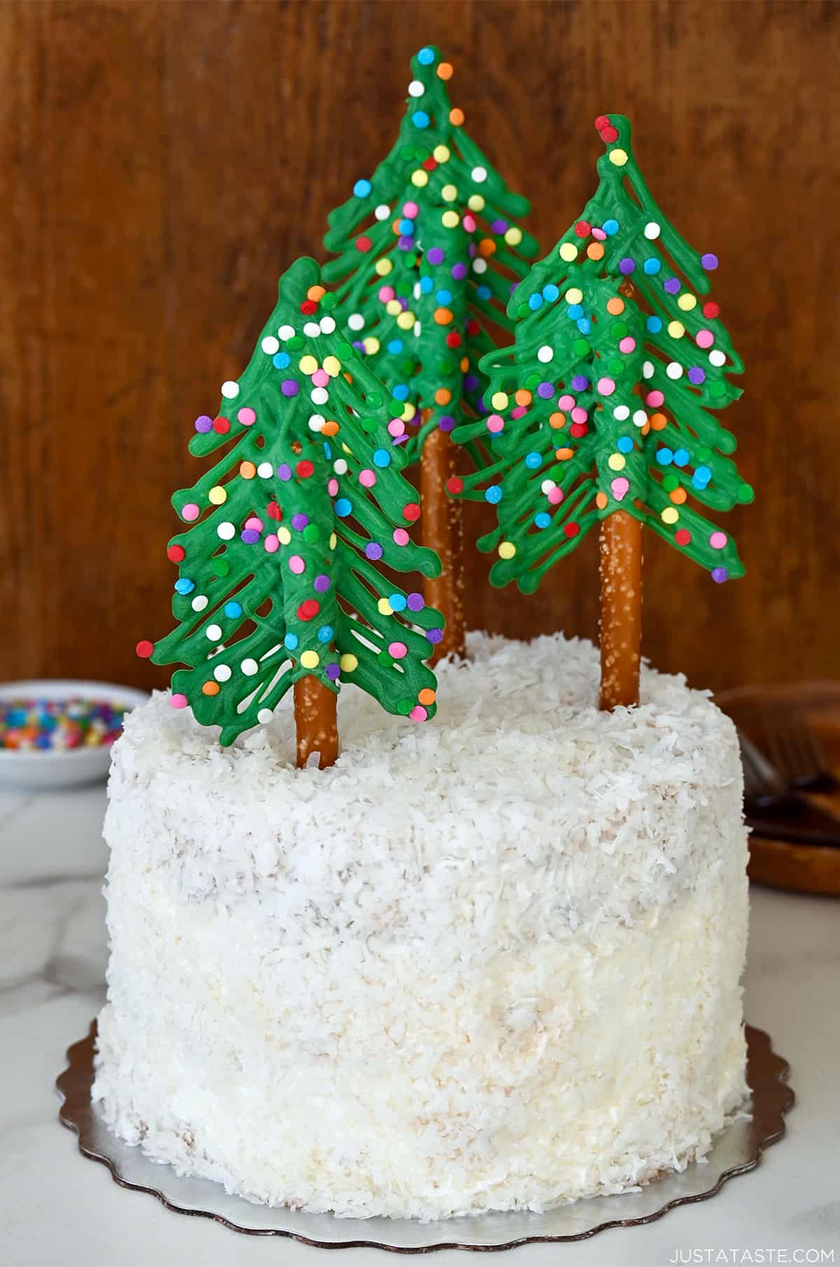 A cake covered in shredded coconut and topped with pretzel rod and candy melt Christmas trees.