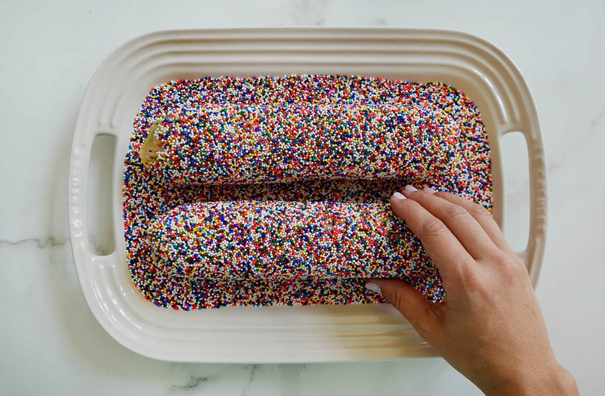 A hand reaches for a rainbow sprinkles-coated cookie dough log on a platter with rainbow sprinkles.