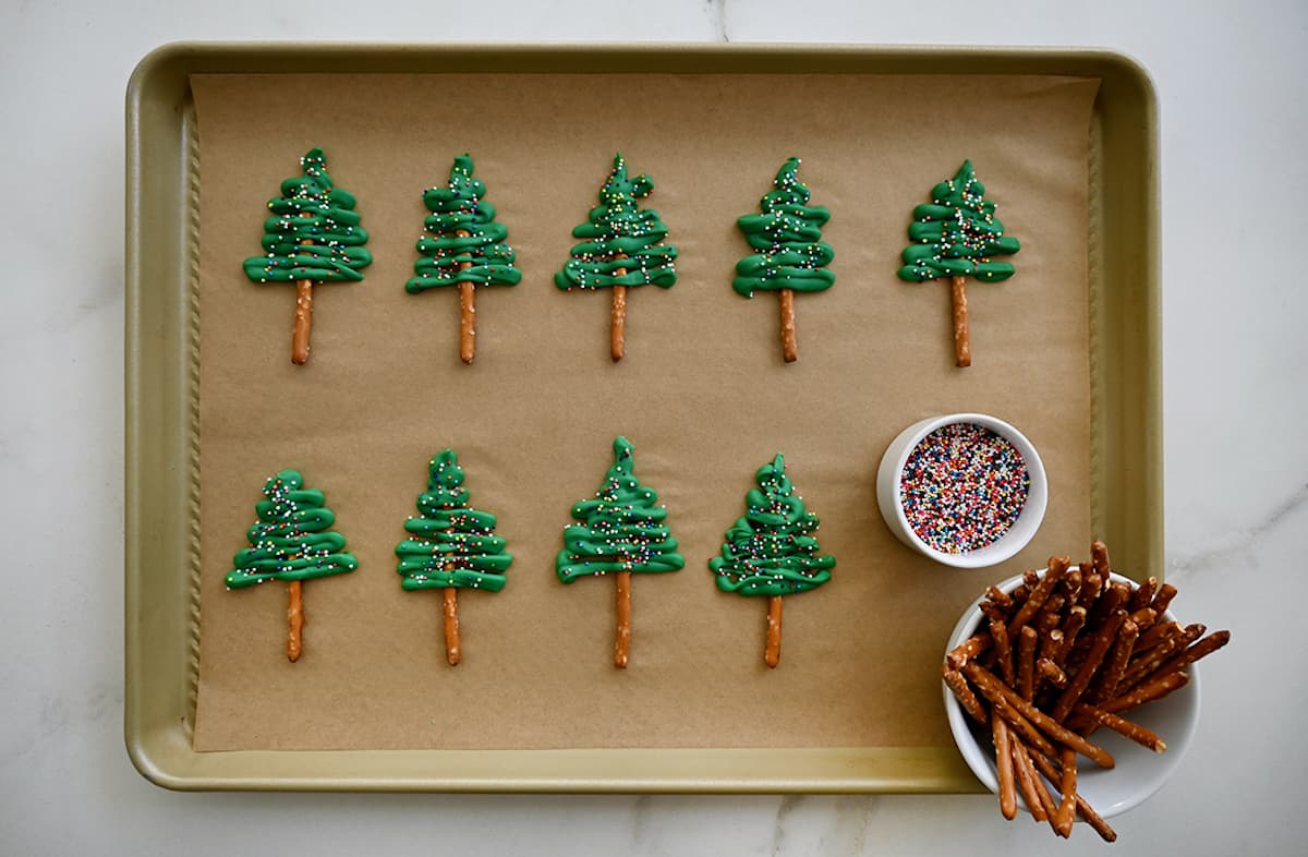 Cupcake toppers made with green candy melts and pretzel sticks on a parchment paper-lined baking sheet next to a small ramekin filled with rainbow sprinkles and a cup filled with pretzel sticks.