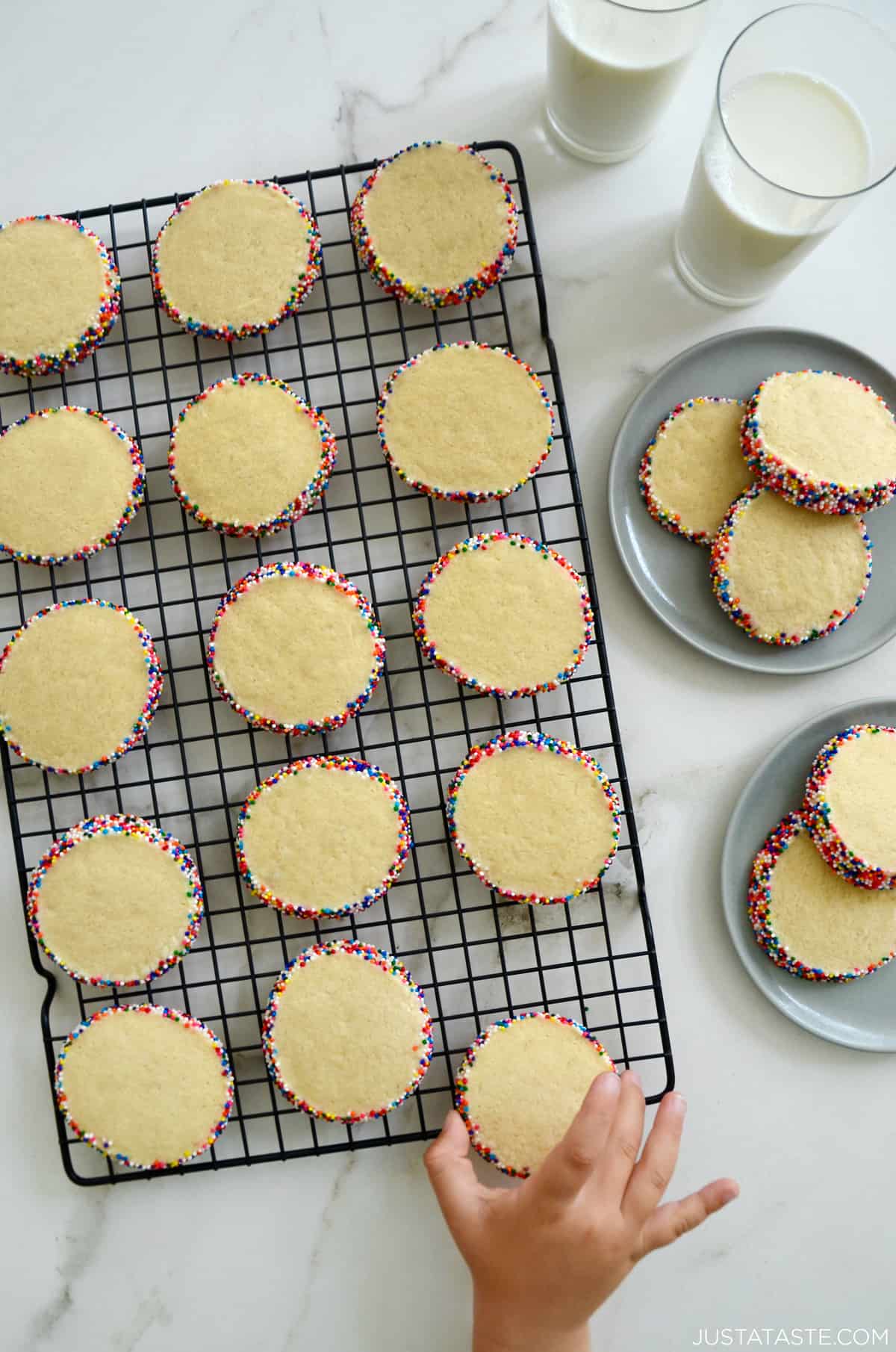 A child's hand reaches for a butter cookie studded with rainbow sprinkles on a wire cooling rack.