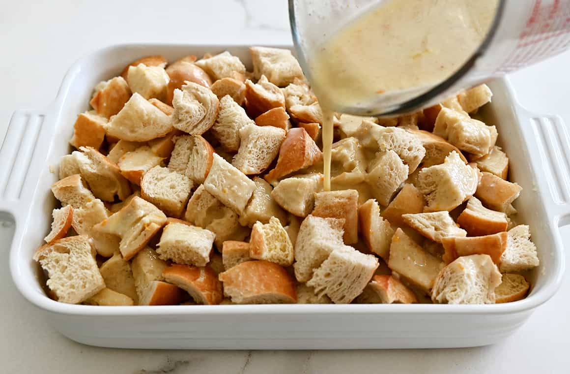 Custard being poured over bread cubes in a white baking dish