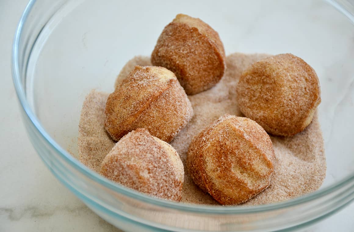 Mini muffins being rolled in a cinnamon-sugar mixture in a clear glass bowl