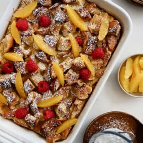 A baking dish containing Overnight French Toast Bake dusted with powdered sugar and topped with orange slices and fresh raspberries
