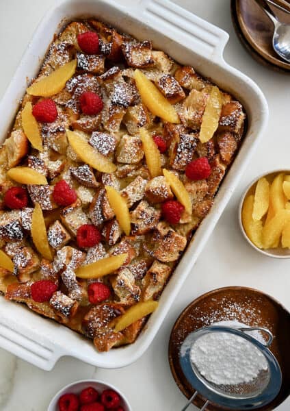 A baking dish containing Overnight French Toast Bake dusted with powdered sugar and topped with orange slices and fresh raspberries