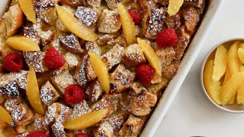 A baking dish containing Overnight French Toast Bake dusted with powdered sugar and topped with orange slices and fresh raspberries