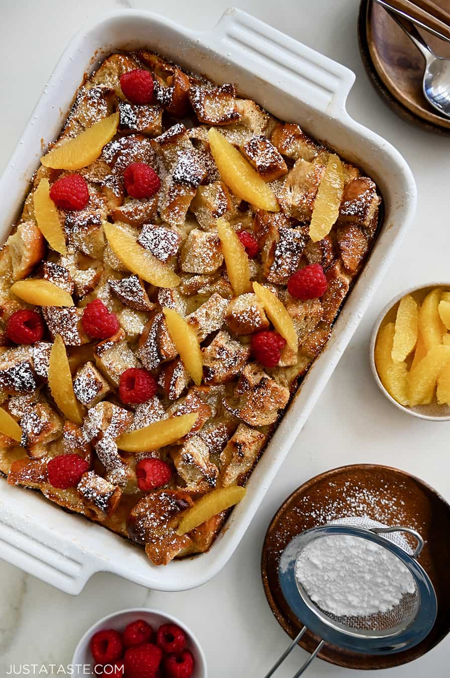 A baking dish containing Overnight French Toast Bake dusted with powdered sugar and topped with orange slices and fresh raspberries