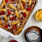 A top-down view of Overnight French Toast Bake in a white baking dish topped with fresh raspberries and orange slices, and dusted with powdered sugar.