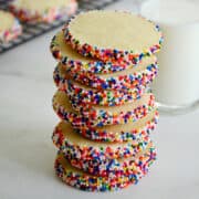 A tall stack of butter cookies studded with rainbow sprinkles in front of a glass filled with milk.